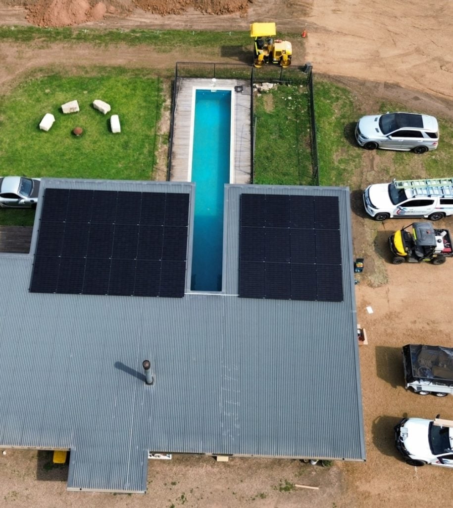 aerial view of a house with solar panels on the roof and a swimming pool in the backyard, surrounded by a dirt road and parked vehicles